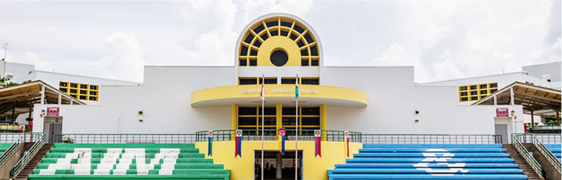 Front view of Tampines Junior College building with yellow facade, arched window, flags, and colorful bleachers.